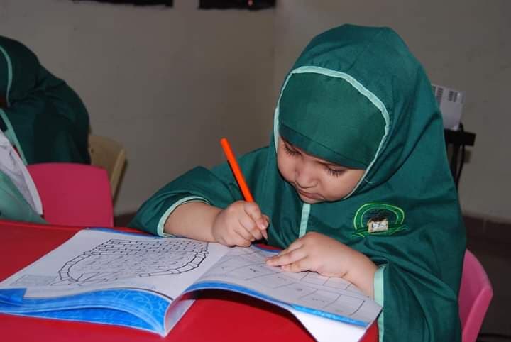 Nursery pupil in green hijab attentively writing in her workbook